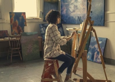 Teenage girl sitting infront of an easel painting with art in the background, highlighting the importance of art mindfulness on stress reduction and headaches in adolescents