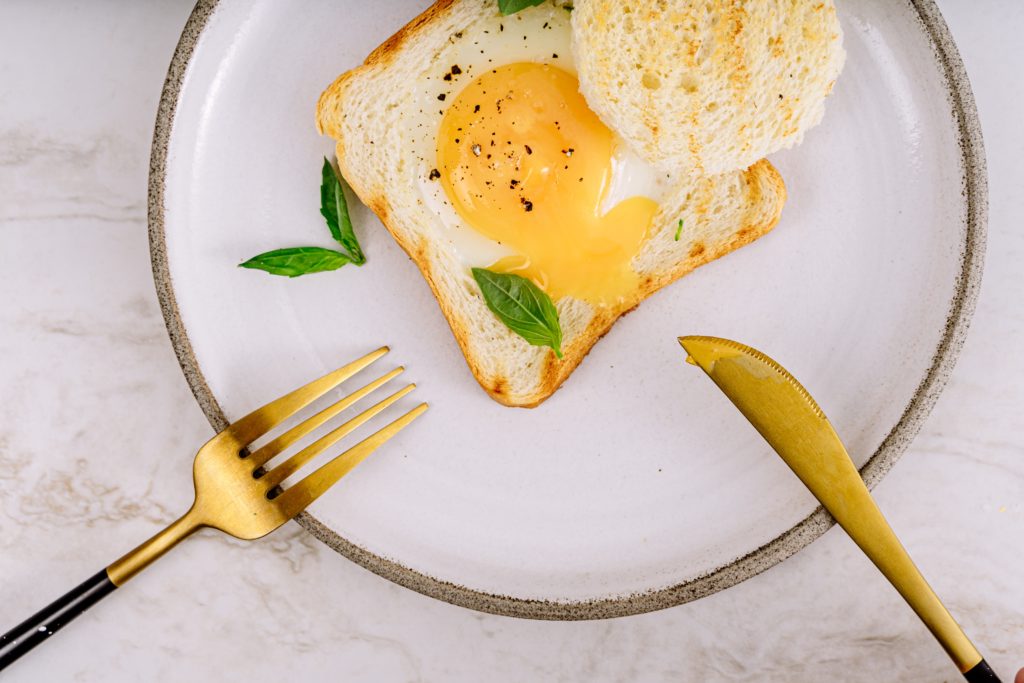 Egg in a hole on white bread sitting on a white plate with a gold rim and gold cutlery resting on the edge of the plate; relating to egg yolks being a good source of Vitamin D.