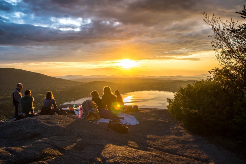 a group watching the sunset on a hike promoting social connection, with yellows and oranges over mountains in the background