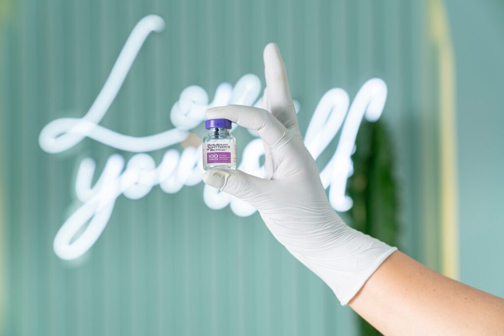 a background neon light that says Love Yourself, with a hand holding a Medical Aesthetics treatment vial