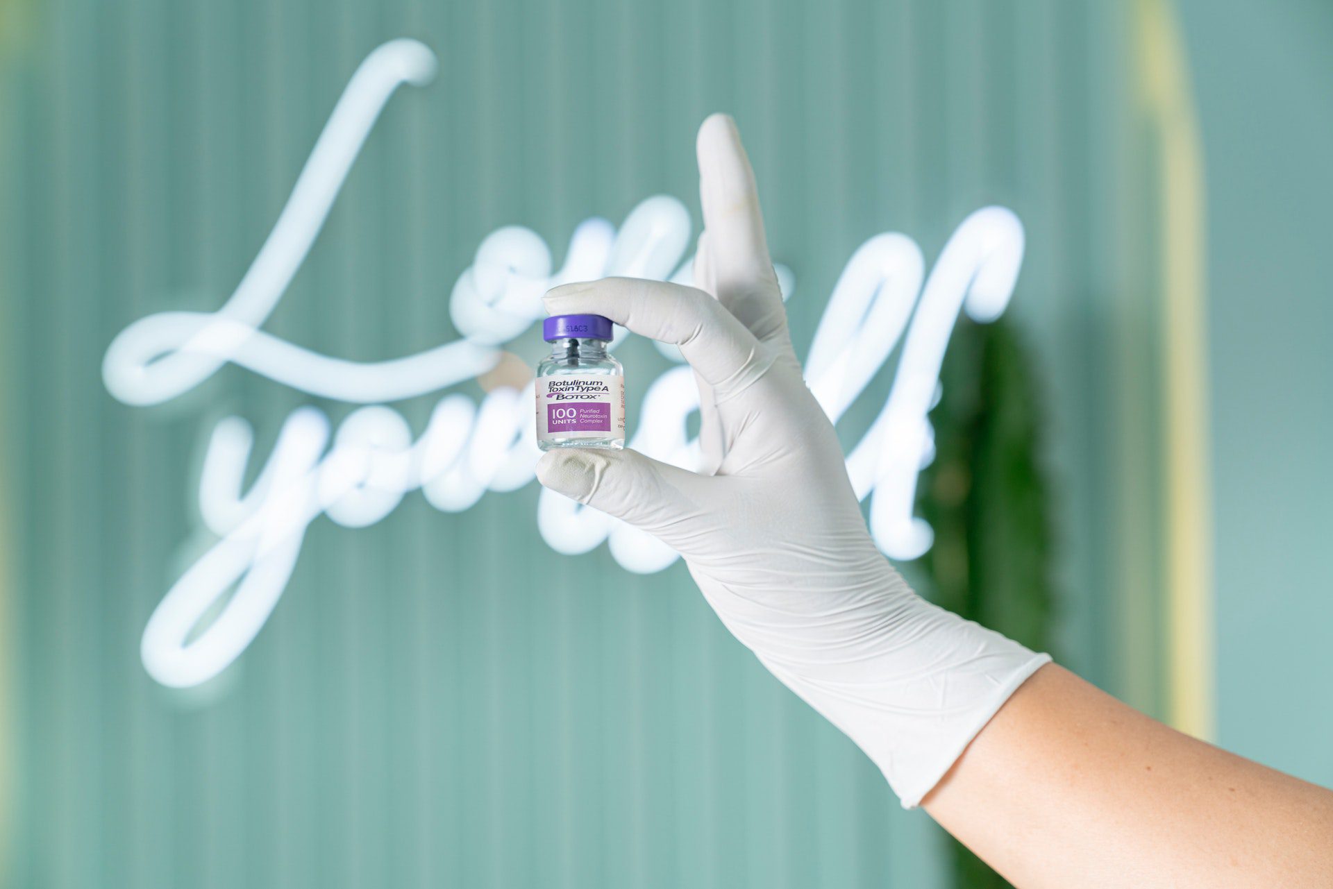 a background neon light that says Love Yourself, with a hand holding a Medical Aesthetics treatment vial