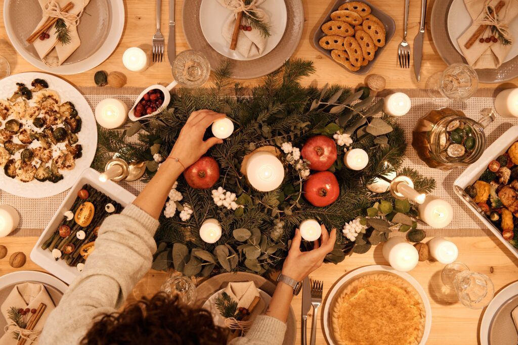 A table set for a festive holiday dinner complete with food, dishes, and glasses, with a person reaching in to place candles candles; highlighting  holiday hacks, such as eating together, that can help relieve stress. 