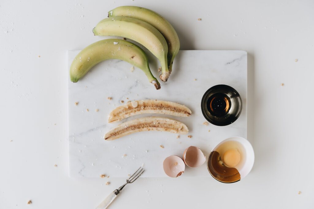 a marble cutting board on top of a countertop with bananas, an egg cracked into a bowl, and a fork. 