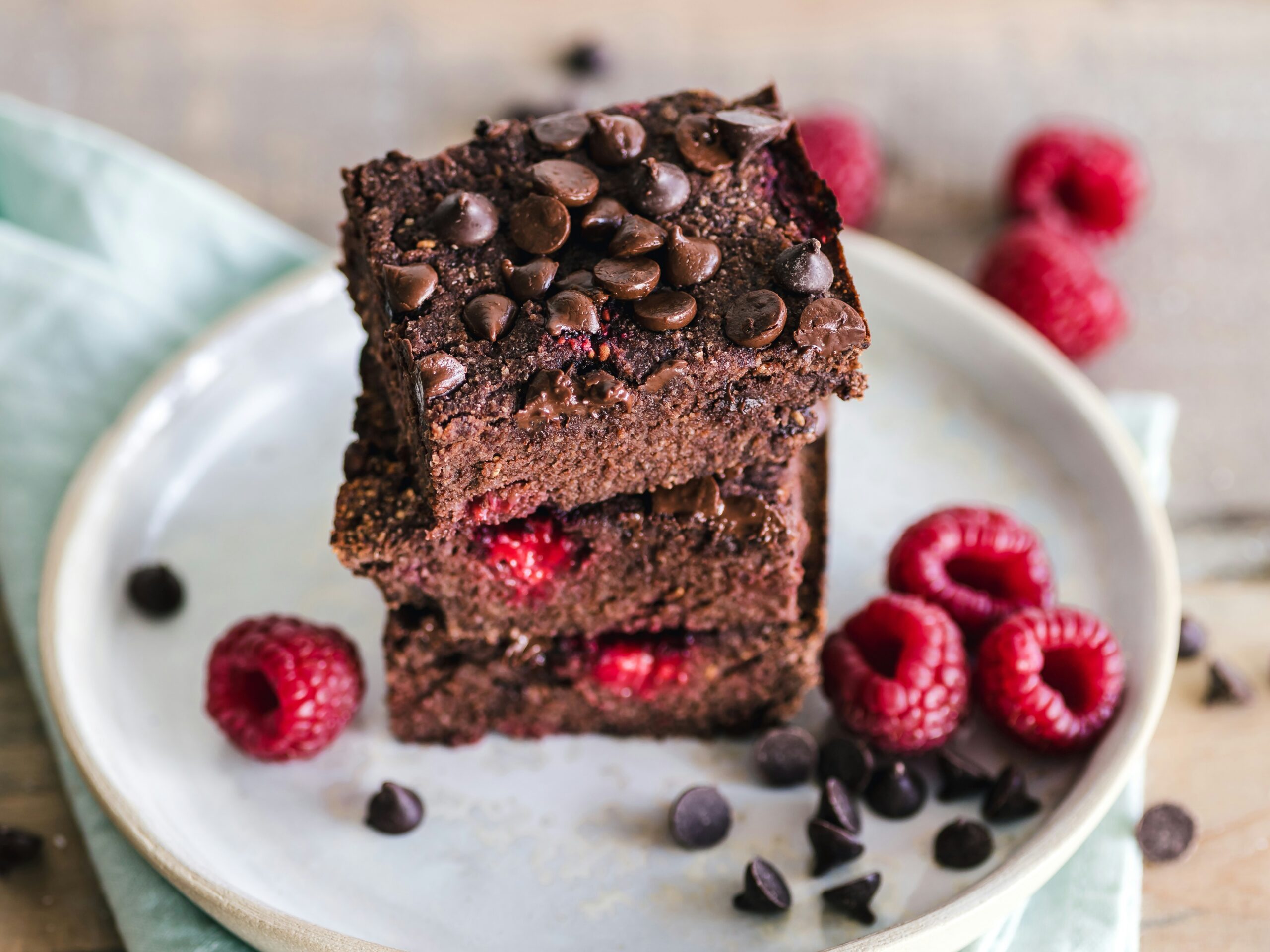 Three raspberry fudge brownies stacked on a white side plate with chocolate chips and fresh raspberries scattered around the plate.