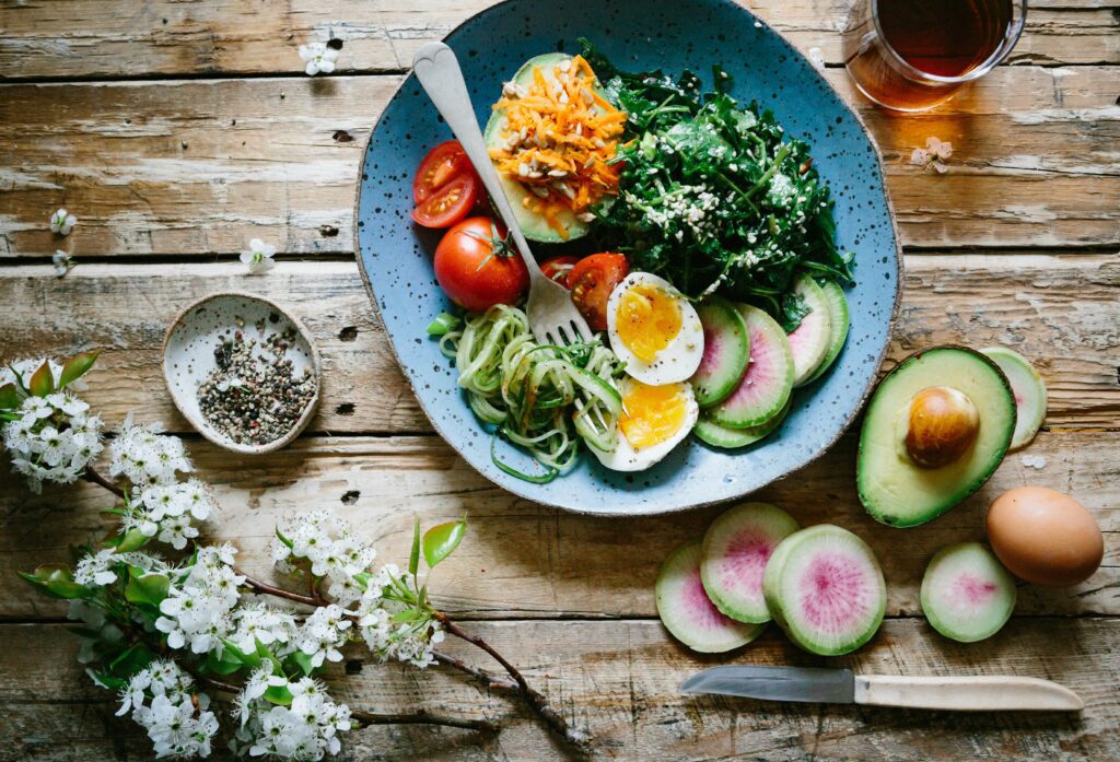 A heart healthy bowl of eggs, figs, avocados, tomatoes, and spinach on a wood table with a sprig of apple blossoms, a knife, and more figs, a half avocado and an egg nearby.